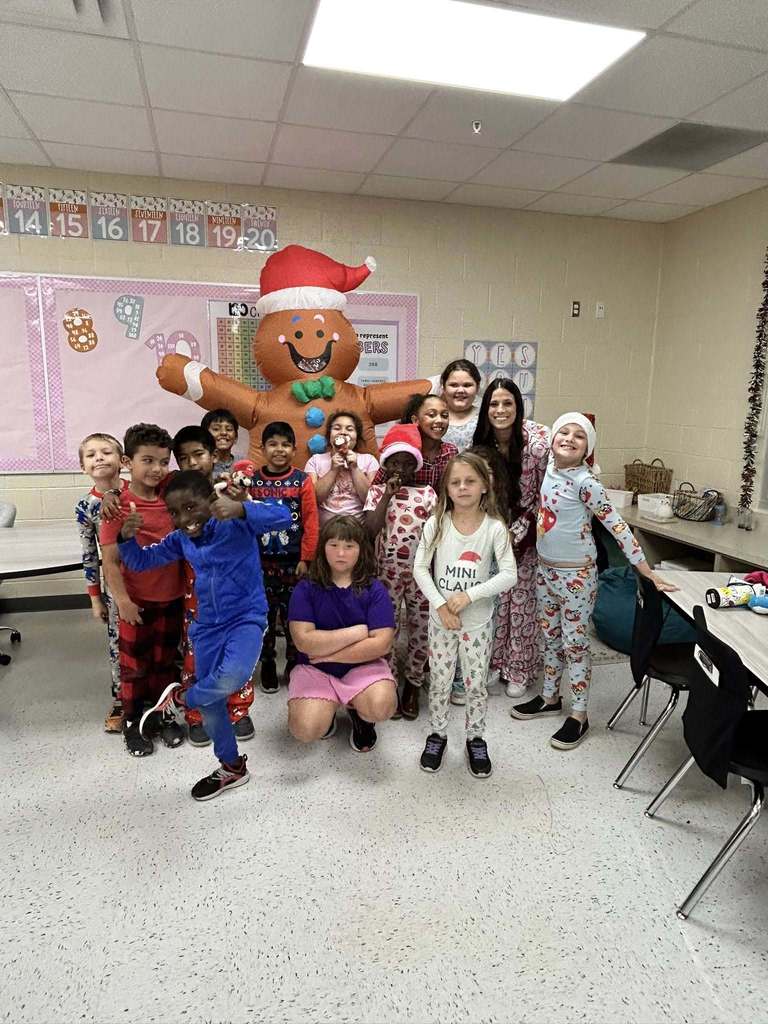 A diverse group of school children and a teacher pose in a classroom with a large inflatable gingerbread man wearing a Santa hat. The children wear festive pajamas, and a boy in a blue tracksuit strikes a playful pose in the foreground.