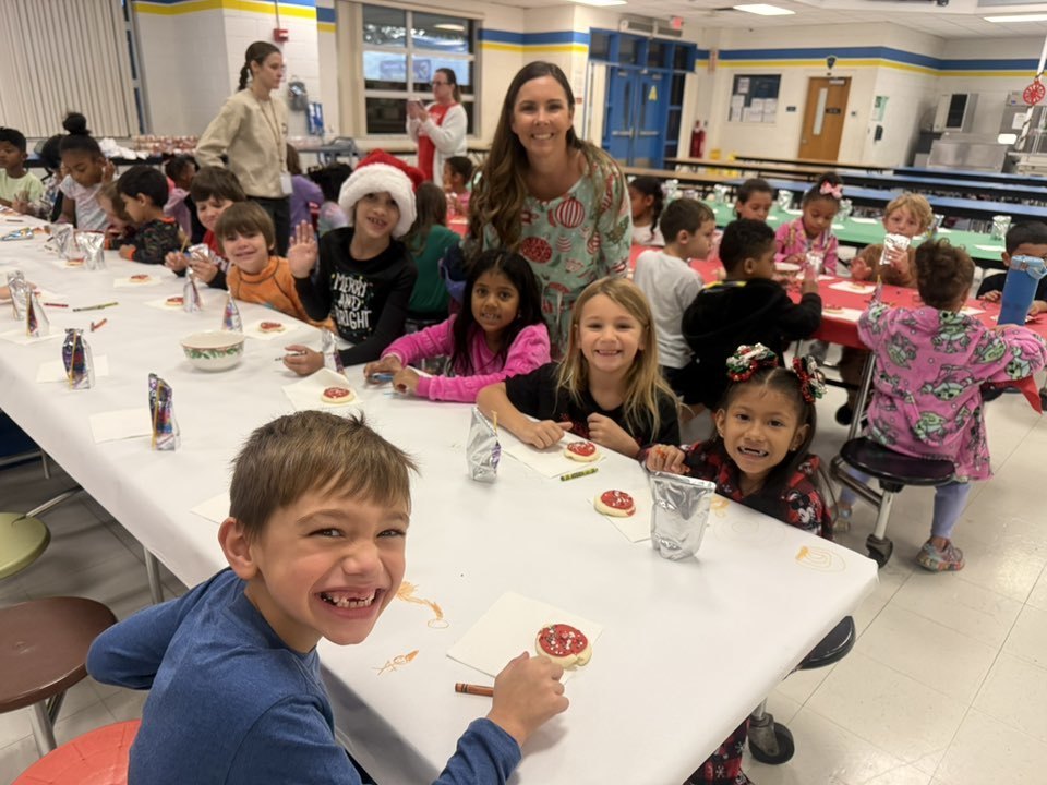A group of elementary students and a teacher sit at a long table in a school cafeteria. The children are decorating holiday cookies and drinking from silver pouches. Several children smile for the camera, including a boy in the foreground with a wide, missing-tooth grin.