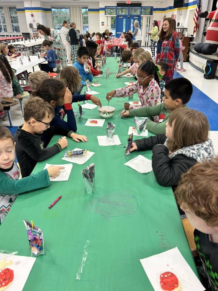Students in pajamas sit along a long green table in a cafeteria, decorating holiday cookies and drawing on the paper table covering. In the background, other students and staff gather near a large inflatable Santa Claus and an American flag.