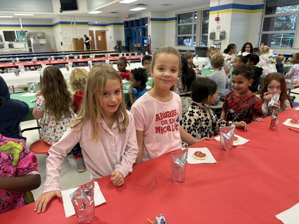 Two young girls stand at a long table covered in red paper in a school cafeteria. One girl wears a pink shirt that says "Always on the Nice List." The table is set with holiday cookies on napkins and silver juice pouches for several seated students.