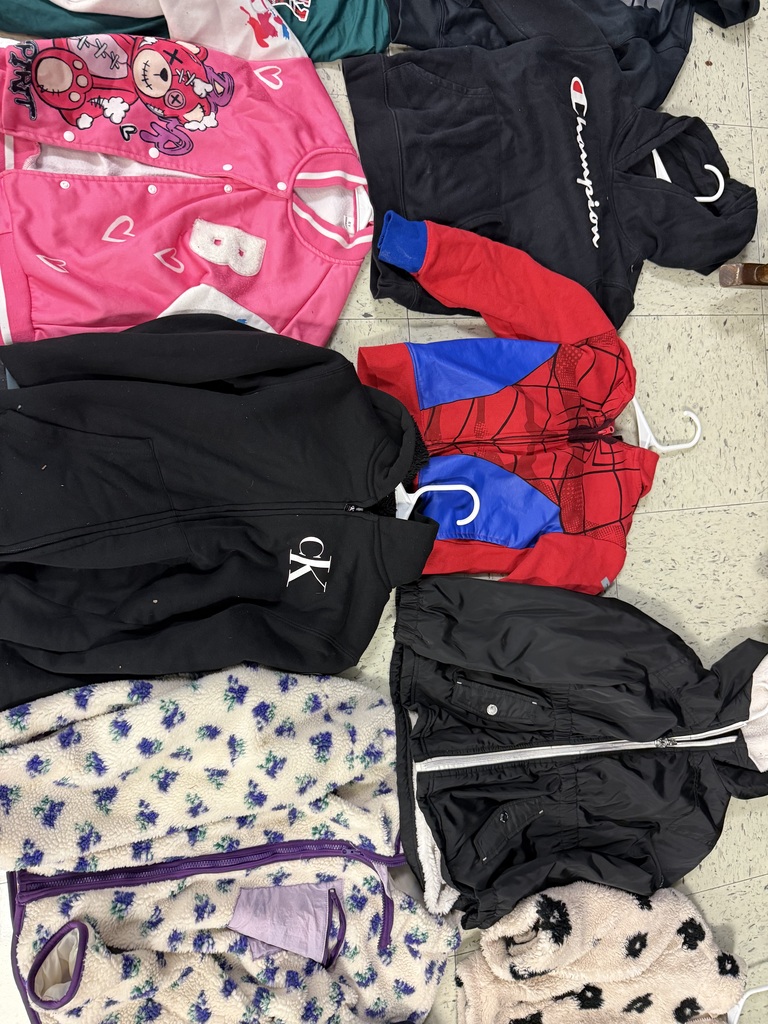 An assortment of children's jackets on a tiled floor, featuring a pink bear varsity jacket, a black Champion hoodie, a Spider-Man hoodie, a black Calvin Klein zip-up, and several patterned fleece jackets.