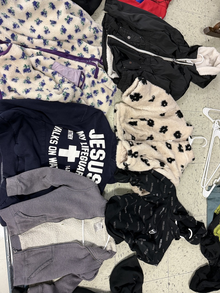 Multiple tops laid out, including a grey sweatshirt, a black cinched hoodie, a Spider-Man mask hoodie, a pink zip-up, a black "Justice Sport" velour jacket, and a green plaid flannel shirt.