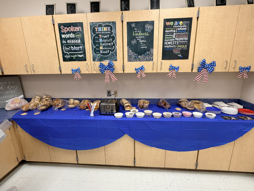 A long table covered with a blue tablecloth displays assorted bagels and containers of spreads arranged in a row, with plates and napkins stacked on the side. Red, white, and blue bow decorations hang above the table, and confetti is scattered across the table, creating a festive setup.