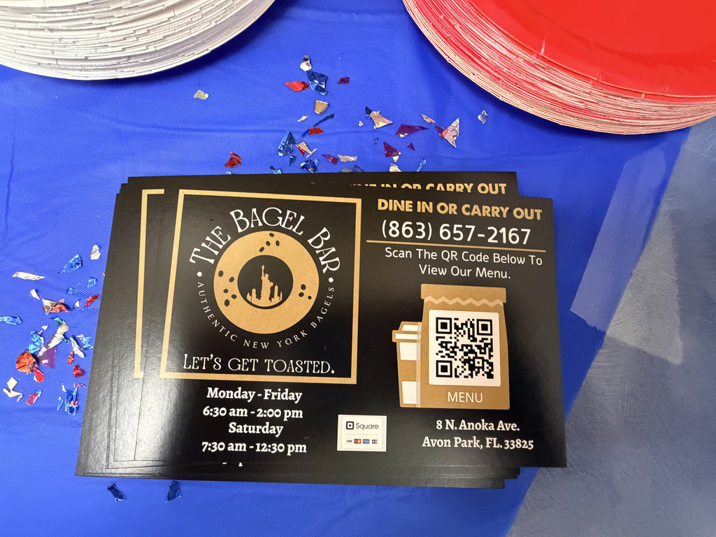 A stack of black and gold flyers for The Bagel Bar sits on a blue tablecloth, surrounded by red, white, and blue plates and scattered confetti. The flyer features The Bagel Bar logo, a QR code for the menu, business hours, phone number, and the slogan “Let’s get toasted.”