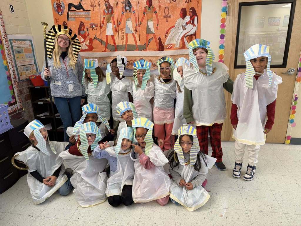 A teacher and group of young children pose in a classroom wearing DIY Ancient Egyptian costumes, including striped paper pharaoh headdresses and white plastic tunics. A large Egyptian mural hangs behind them. The students are smiling and gathered for a group photo.