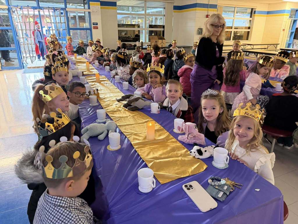 A large group of young children wearing gold paper crowns sit at long tables in a bright cafeteria. The tables feature purple tablecloths and gold runners with white mugs and stuffed animals. An adult stands in the background as the children smile for a group photo.