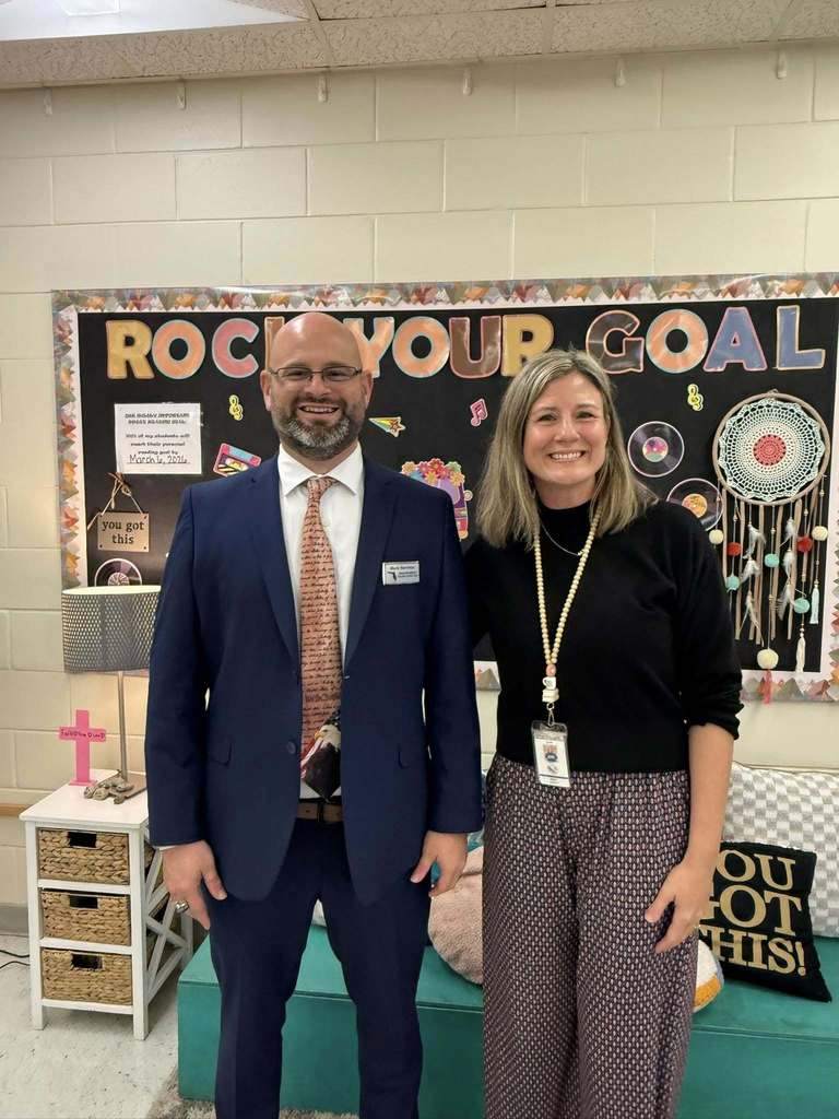 A man in a blue suit and patriotic tie stands next to a woman in a black sweater and patterned pants. They are smiling in a classroom in front of a bulletin board that reads "ROCK YOUR GOAL." The room includes a small white side table and a teal bench.
