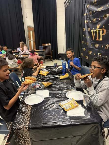  Several children sit at a long table covered in black plastic, eating pizza and snacks. A boy in the foreground bites into a slice while another holds his up. Behind them, a "VIP" backdrop hangs against dark curtains, and other students eat at a table in the background.