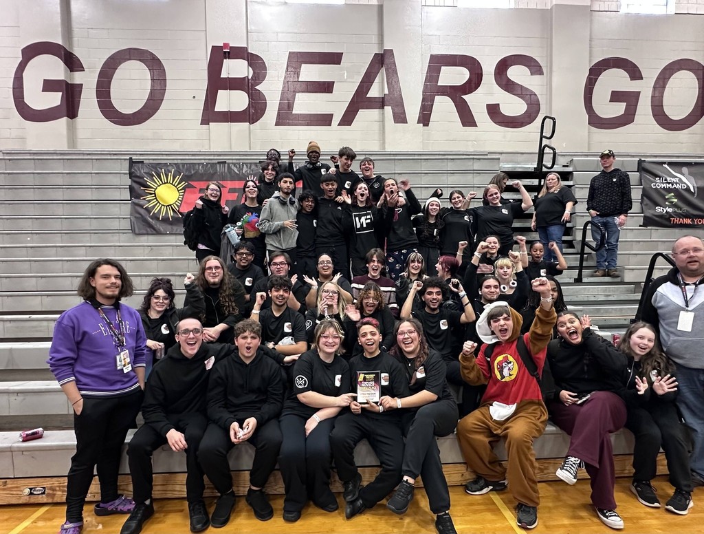 The same group of students sits more composed on the bleachers for a formal photo. They are mostly dressed in black, with a student in the center holding a third-place plaque. The "GO BEARS GO" wall and "FFCC" banner remain visible in the background as they smile for the camera.