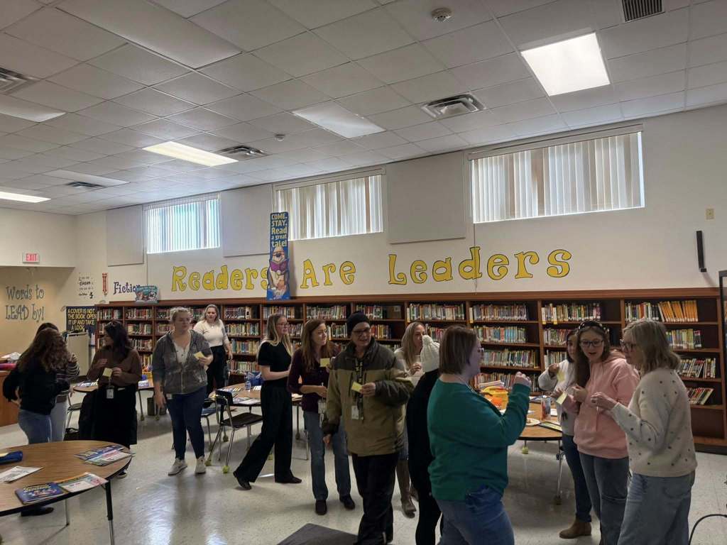  A brightly lit school library features a group of adults gathered near circular tables for an activity. The background shows bookshelves and a wall with the yellow text "Readers Are Leaders." Some individuals hold yellow sticky notes, while others converse in small groups under fluorescent ceiling lights.