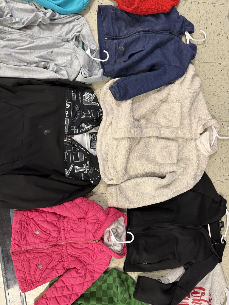 A pile of children’s jackets on hangers, featuring a bright pink quilted puffer jacket, a cream sherpa coat, a black jacket with a patterned lining, and several navy and gray zip-ups.