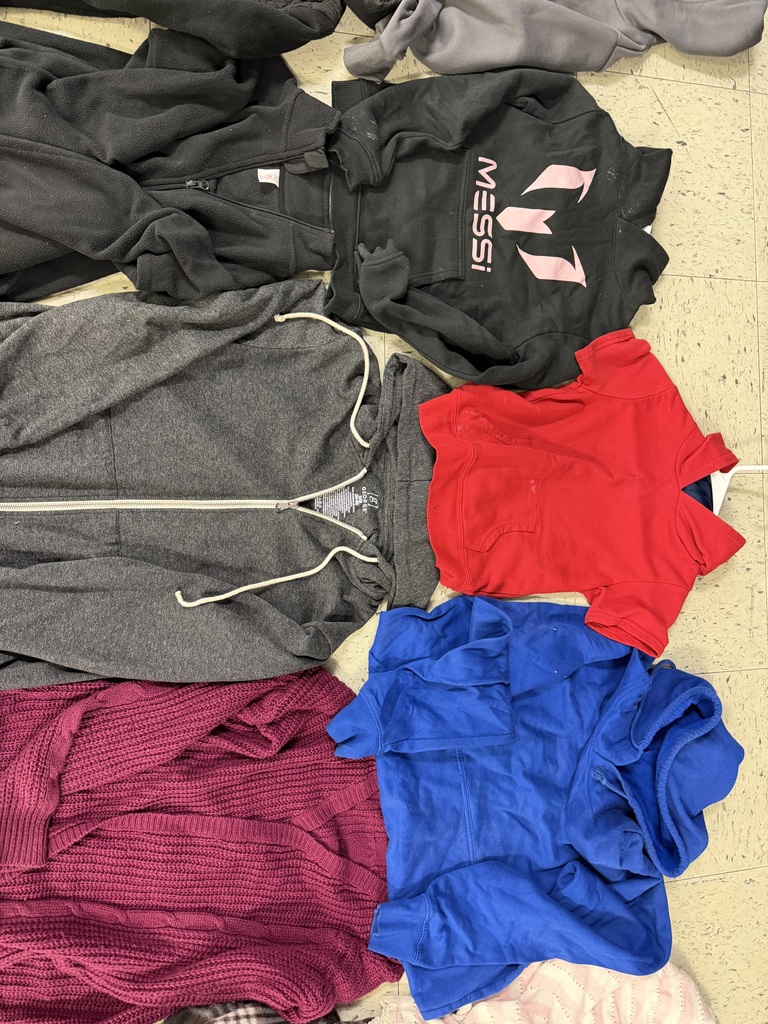Various sweatshirts and sweaters on a floor, including a black "MESSI" hoodie with pink lettering, a gray zip-up, a bright red hoodie, a royal blue hoodie, and a maroon knit cardigan.