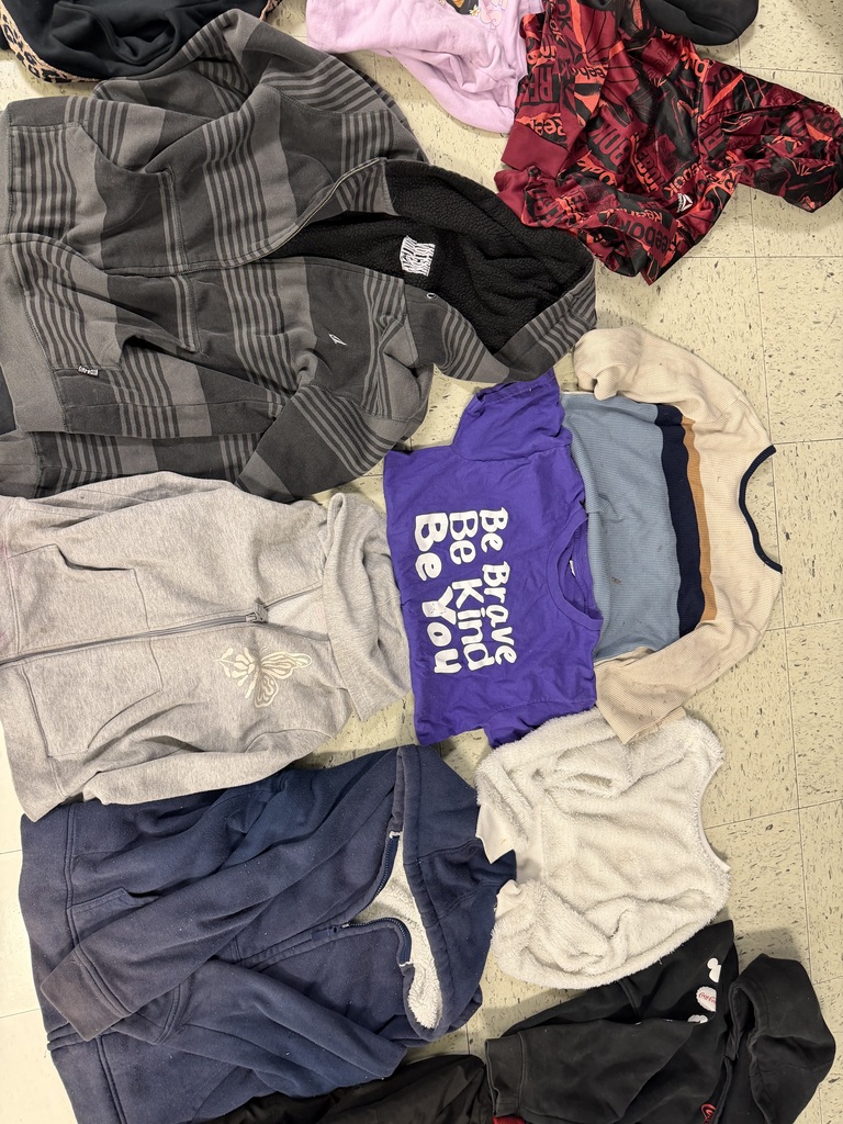 Various children’s clothing items scattered on a floor, featuring a large gray-striped fleece-lined hoodie, a purple "Be Brave Be Kind Be You" t-shirt, a white fuzzy sweater, and several other colorful hoodies and knit tops.