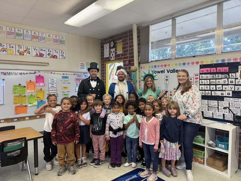 A classroom group photo shows about 20 young students standing with four adults. Three adults are in costume: Abraham Lincoln, George Washington, and the Statue of Liberty. They are gathered in a bright classroom featuring educational posters like "Vowel Valley" and an alphabet chart on the walls.