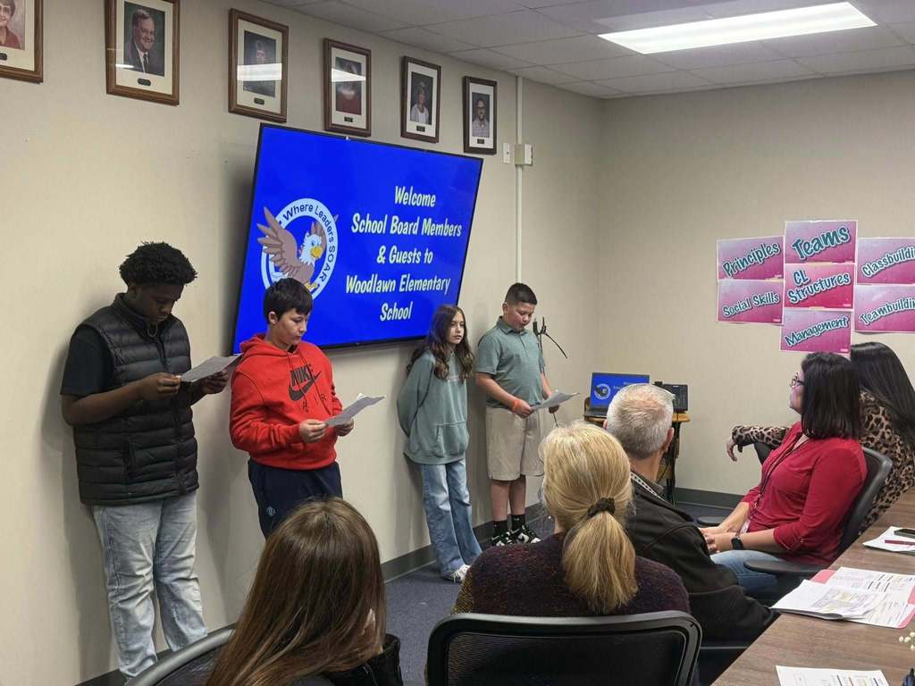  Gemini said Four students stand in a room, some reading from papers, presenting to a seated group of adults. A large monitor behind them displays "Welcome School Board Members & Guests to Woodlawn Elementary School." Framed portraits and educational posters about "Teams" and "Social Skills" decorate the walls.