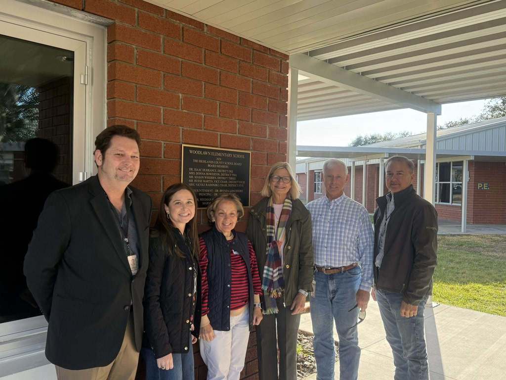 Six adults stand outside a brick building next to a commemorative plaque for Woodlawn Elementary School. The plaque lists "The Highlands County School Board" members for 2024. The group is smiling, positioned under a covered walkway with additional school buildings in the background.