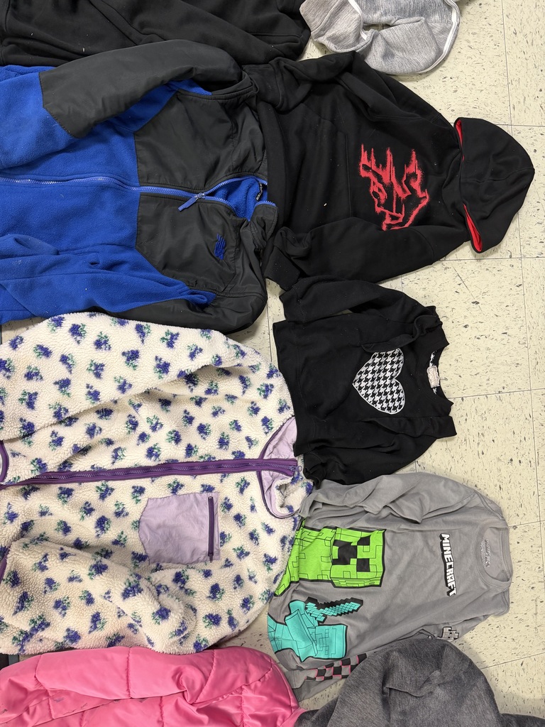 A group of children's clothing. Notable items include a white fleece jacket with a purple floral pattern, a black hoodie with a red graphic, a grey Minecraft t-shirt, and a bright pink puffer jacket.