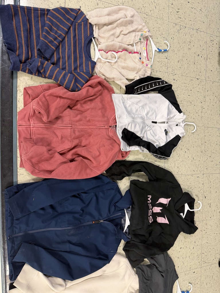 Six items of clothing, some on white hangers. Includes a navy and orange striped sweater, a tan knit cardigan with colorful pom-poms, a red zip-up hoodie, and a black and white Nike track jacket.