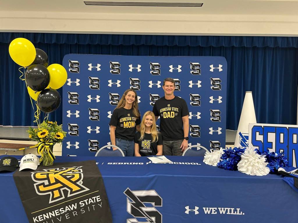 A student-athlete sits at a blue signing table flanked by her parents, who wear "Kennesaw State Mom" and "Dad" shirts. They are positioned in front of a blue Kennesaw State University backdrop with yellow and black balloons, sunflowers, and school branded apparel on the table.
