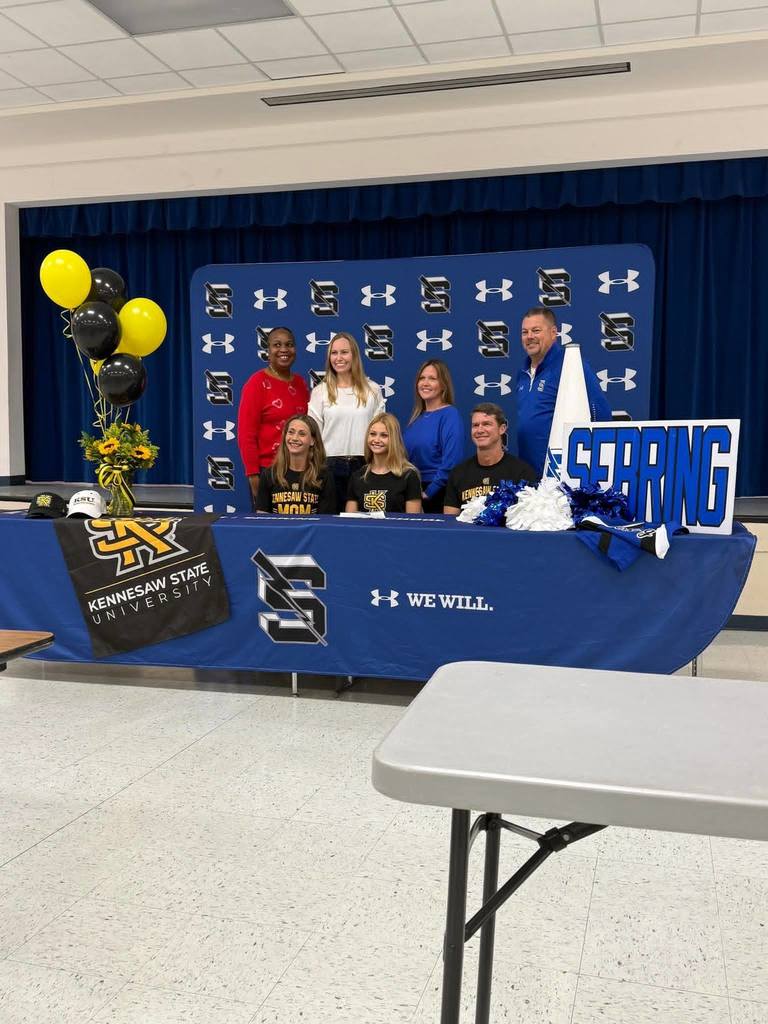 A group photo at a college signing event features the student and her parents seated at a blue table, with four adults standing behind them. The setting includes Kennesaw State University branding, blue pom-poms, a "Sebring" sign, and a backdrop displaying the school's logo and the Under Armour "We Will" slogan.