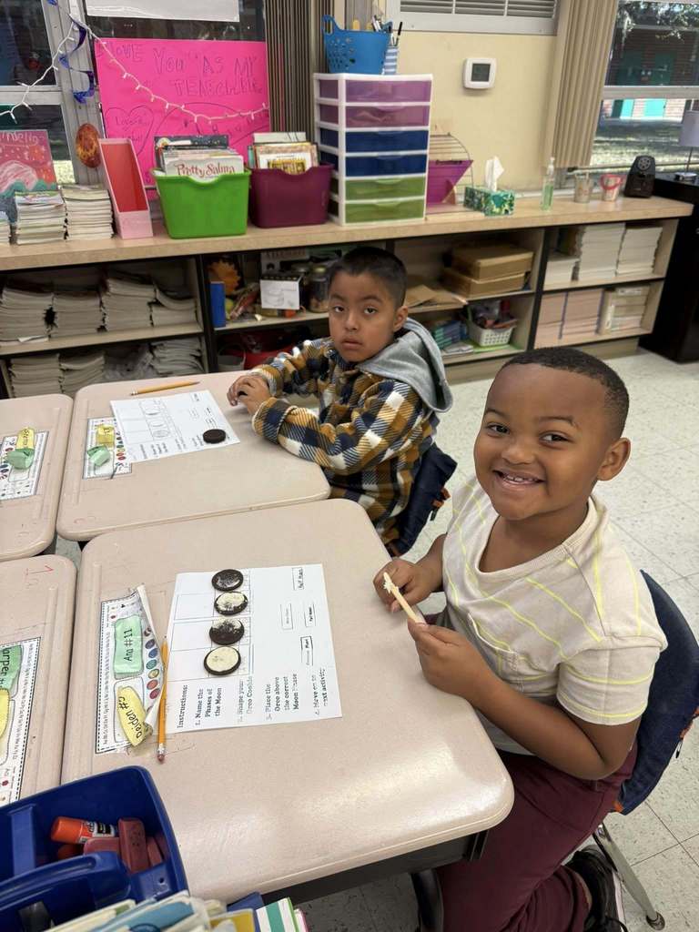 Two young students sit at classroom desks, participating in a science activity. They are using Oreo cookies to represent the phases of the moon on a worksheet. One student smiles at the camera while holding a wooden stick, with organized classroom shelves and supplies in the background.
