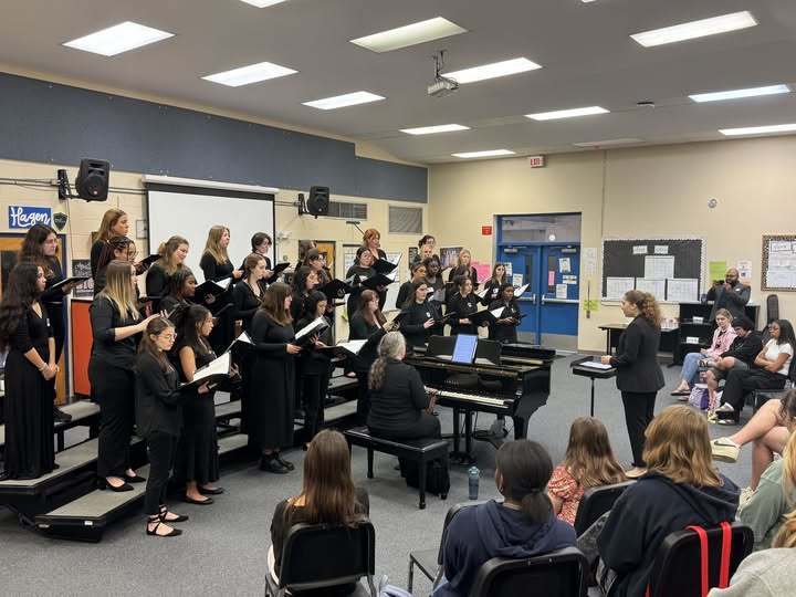 An all-female choir in black formal attire performs on risers in a classroom. A conductor stands before them, and a pianist plays a grand piano. An audience sits in the foreground. The room features fluorescent lighting, a projector screen, and educational posters on the walls.