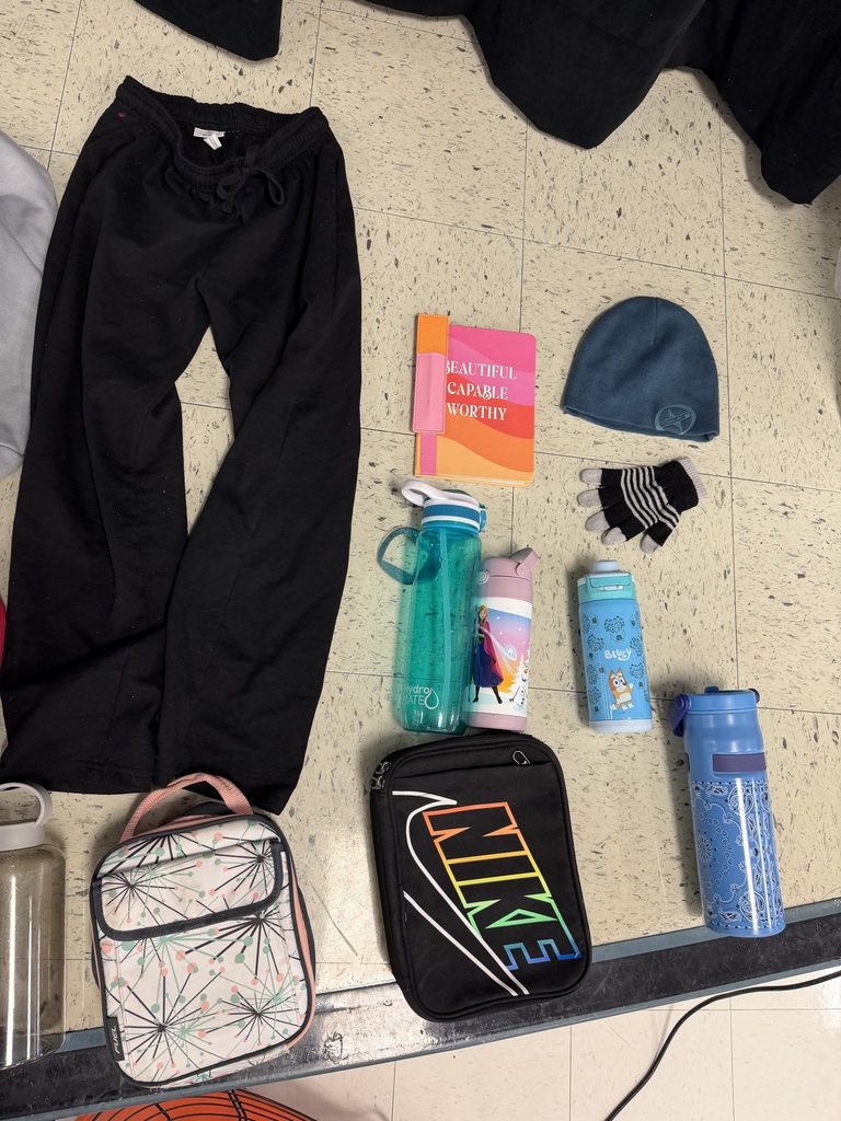 A pair of black sweatpants laid out next to various accessories: a "Beautiful Capable Worthy" notebook, a teal beanie, striped gloves, a Nike lunch bag, a patterned lunch box, and four different reusable water bottles including one featuring "Frozen" characters.