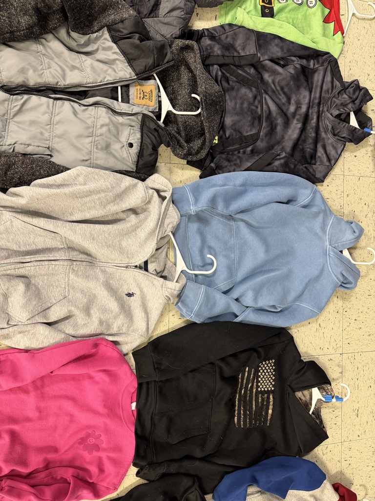 A spread of five garments: a grey quilted zip-up jacket, a dark grey patterned hoodie, a light blue hoodie, a black hoodie featuring a camouflage American flag, and a bright pink sweatshirt with a subtle flower graphic on the bottom left corner.