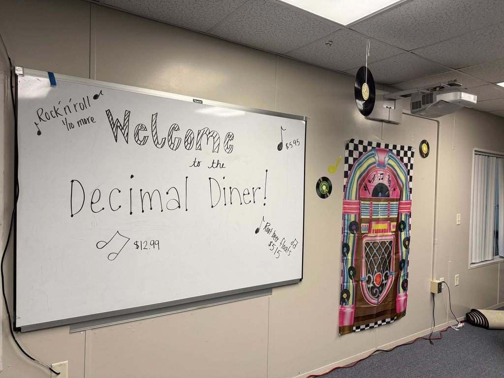  Gemini said A whiteboard reads "Welcome to the Decimal Diner!" in a 1950s rock-and-roll theme. It features prices like $12.99 and $5.15 alongside musical notes. A jukebox poster, hanging vinyl records, and a ceiling-mounted projector decorate the surrounding classroom wall, creating an immersive educational setting.