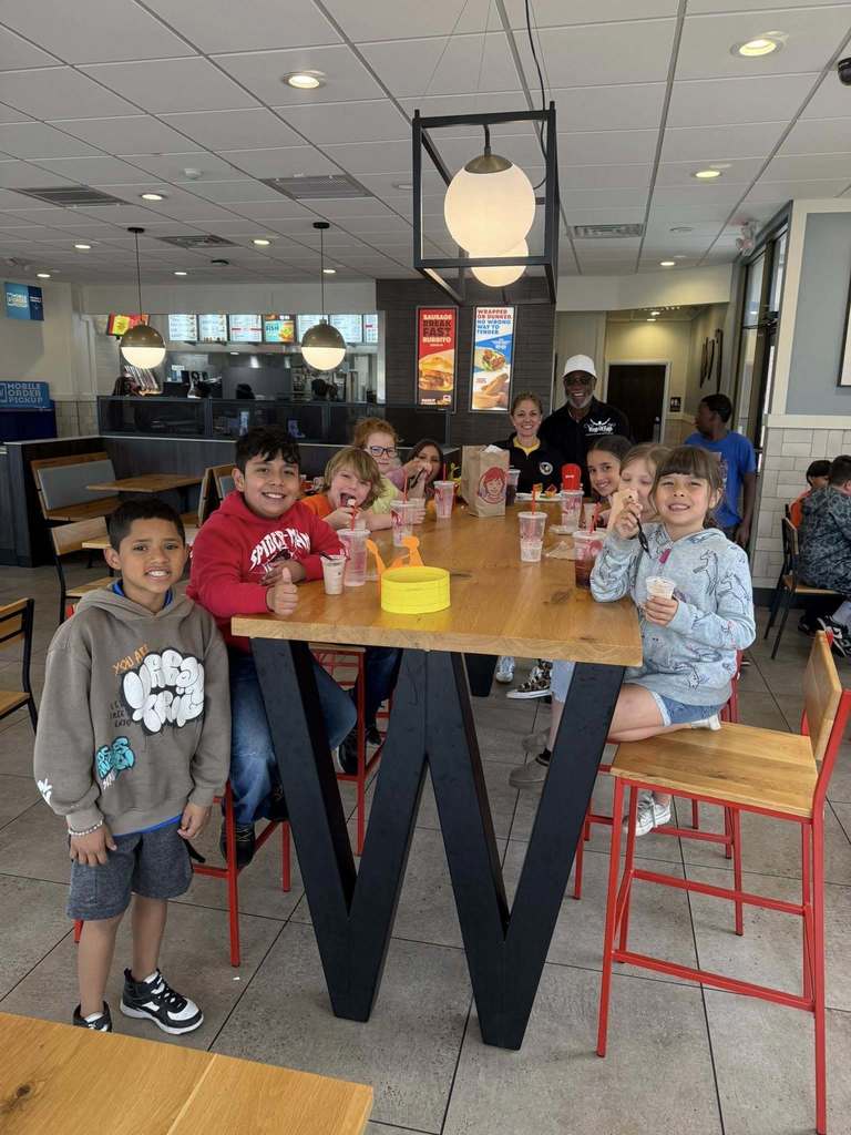 A group of smiling children and two adults sit and stand around a large, "W"-shaped wooden table inside a Wendy’s restaurant. They are enjoying Frostys and meals. The bright, modern interior features hanging globe lights, red-framed chairs, and a visible service counter in the background.