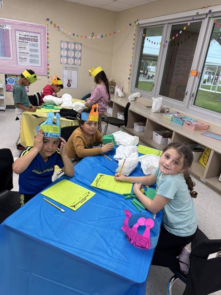 Gemini said Several young students sit at a blue table in a bright classroom, wearing colorful paper crowns. They are working on math activities using yellow worksheets and green blocks. Educational posters and windows line the walls, creating a focused and cheerful learning environment.