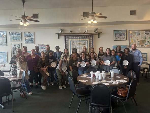 A large group of diverse adults poses indoors, many holding decorated white plates. They are gathered in a room with framed racing posters, a quilt on the wall, and ceiling fans. A round table in the foreground holds white cups and scattered papers. The atmosphere is casual and celebratory.