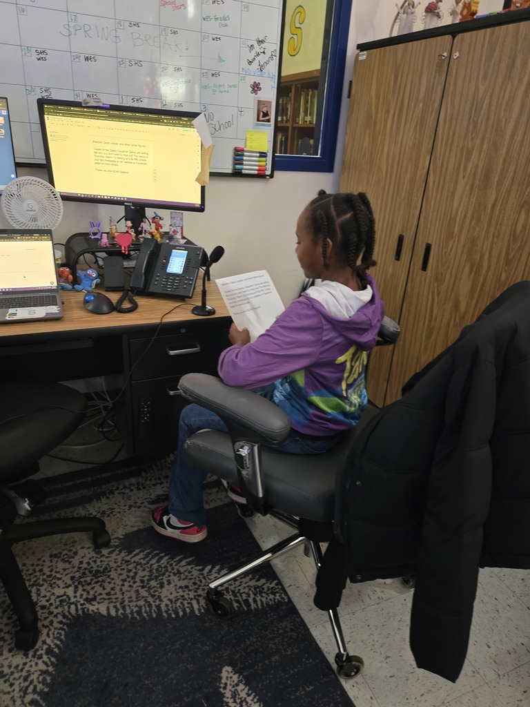 A young girl with braided hair and a purple jacket sits in a black swivel chair at a desk. She is holding a paper and looking toward a computer monitor and a microphone. A white calendar board is visible on the wall behind her.