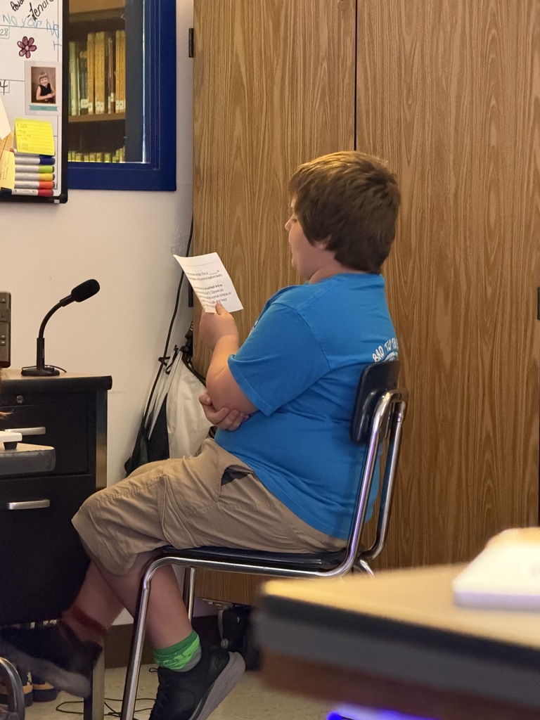 A young boy with brown hair, wearing a blue t-shirt and khaki shorts, sits in a chair holding a piece of paper. He appears to be practicing or reading aloud. A desk with a microphone and a large wooden cabinet are behind him.