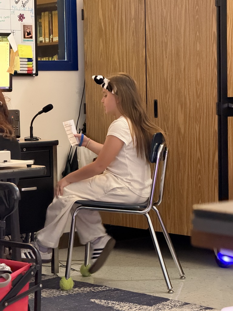 A young girl with long brown hair and a patterned headband sits in a metal chair. She is wearing a white outfit and reading from a small piece of paper. She is positioned in an office-like setting in front of a desk with a microphone.