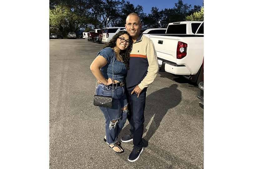 A man and woman pose smiling in a paved parking lot at dusk. The woman wears a blue ruffled top, ripped jeans, and a crossbody bag. The man wears a color-blocked pullover and dark pants. White pickup trucks and trees are visible in the background under a blue sky.