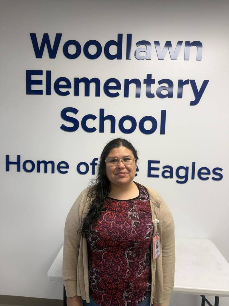 Gemini said A woman with glasses and long dark hair stands in front of a white wall featuring blue text that reads "Woodlawn Elementary School Home of the Eagles." She wears a tan cardigan over a patterned maroon shirt and an ID badge. A white table is visible behind her.