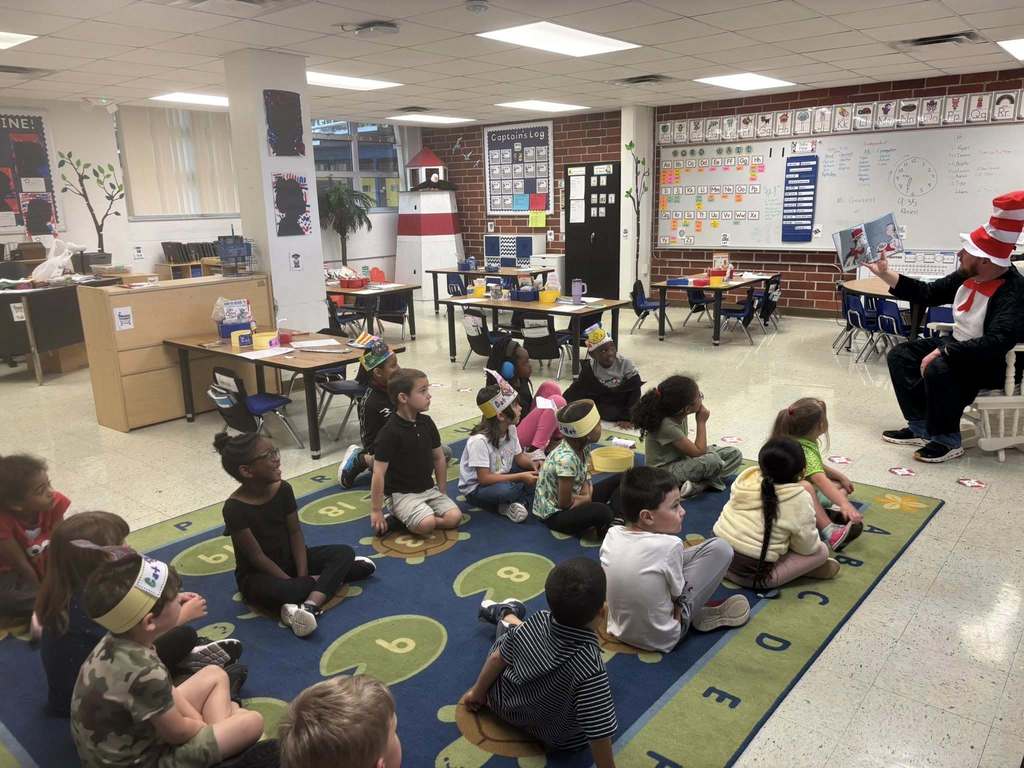 An adult dressed as the Cat in the Hat reads to a group of approximately fifteen young children in a vibrant classroom. The children sit cross-legged on a large, colorful alphabet rug. The room features brick walls, student desks, and educational posters, creating an engaging learning environment.