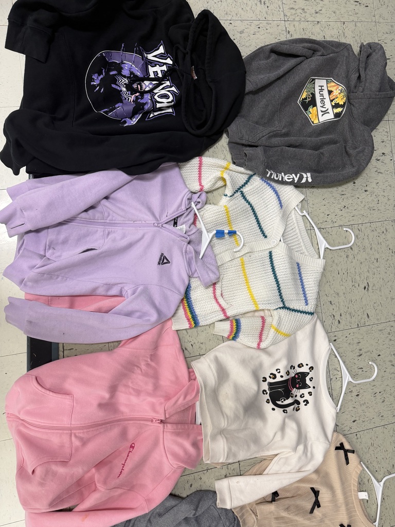 A collection of children's upper-wear on a tiled floor, including a black "Venom" hoodie, a charcoal Hurley hoodie, a lavender Reebok zip-up, a striped white knit cardigan, a pink Champion hoodie, a white sweatshirt with a cat graphic, and a beige ribbed top with black bows.