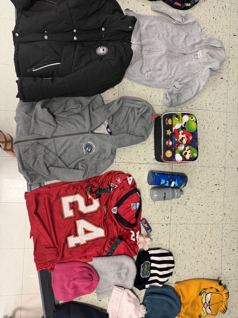 A flat lay featuring a heavy black puffer coat, a grey zip-up hoodie, a red #24 sports jersey, a Mario-themed lunch box, two water bottles, and several colorful beanies (pink, grey, striped, blue, and a yellow Garfield hat).