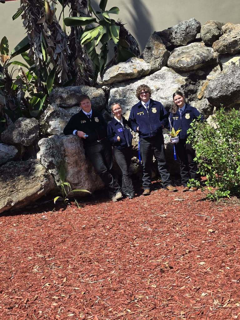 Four students in navy blue FFA jackets and dark trousers stand in a line against a large rock wall. They are positioned outdoors on red wood mulch next to tropical greenery. The group is smiling, and the student on the far right holds a small potted plant.