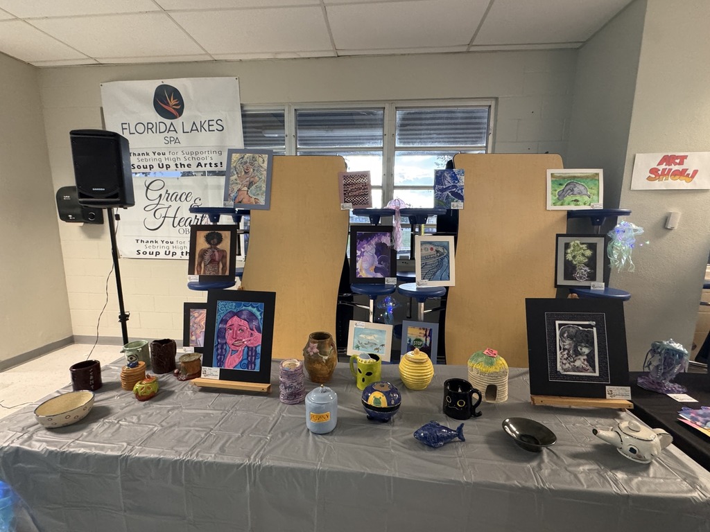Gemini said An art display features a variety of student works on a grey-clothed table. Items include ceramic mugs, bowls, and jars alongside framed colorful drawings and paintings. A banner for "Soup Up the Arts" hangs in the background near wooden partitions and a speaker.