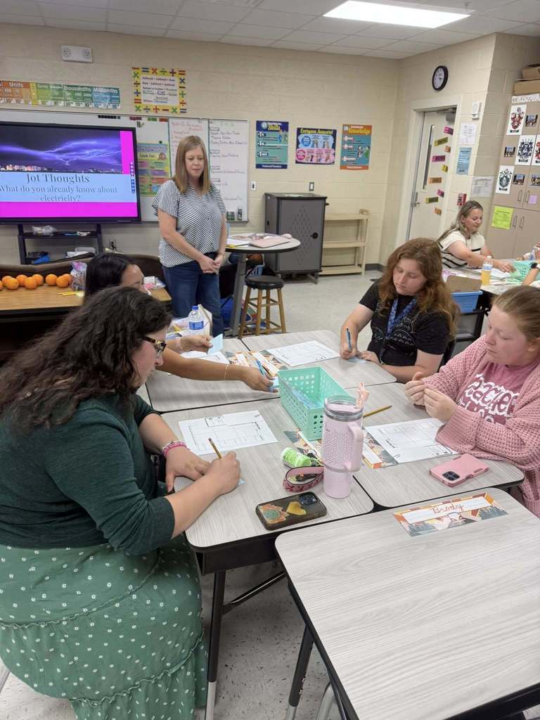 Gemini said A group of adults sits at clustered desks in a classroom, participating in a "Jot Thoughts" activity about electricity shown on a large screen. They are writing on papers and sticky notes. A teacher stands nearby observing, while colorful educational posters and math charts decorate the walls.