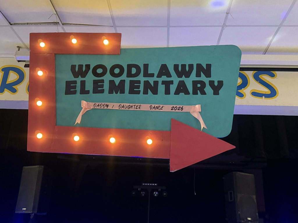 A retro-style marquee sign hangs indoors, featuring a teal background with "WOODLAWN ELEMENTARY" in bold black letters. A pink ribbon below reads "DADDY / DAUGHTER DANCE 2026." The sign is framed by a red L-shaped border with glowing marquee lights and a large red arrow pointing right.