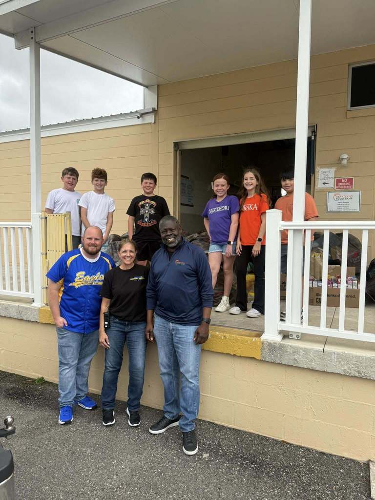 Gemini said A group of nine people—three adults and six children—pose outside the Heartland Food Bank. The adults stand on the ground in front of a loading dock, while the children stand behind them on the elevated platform near stacked boxes of food. Everyone is smiling in a casual, outdoor setting.