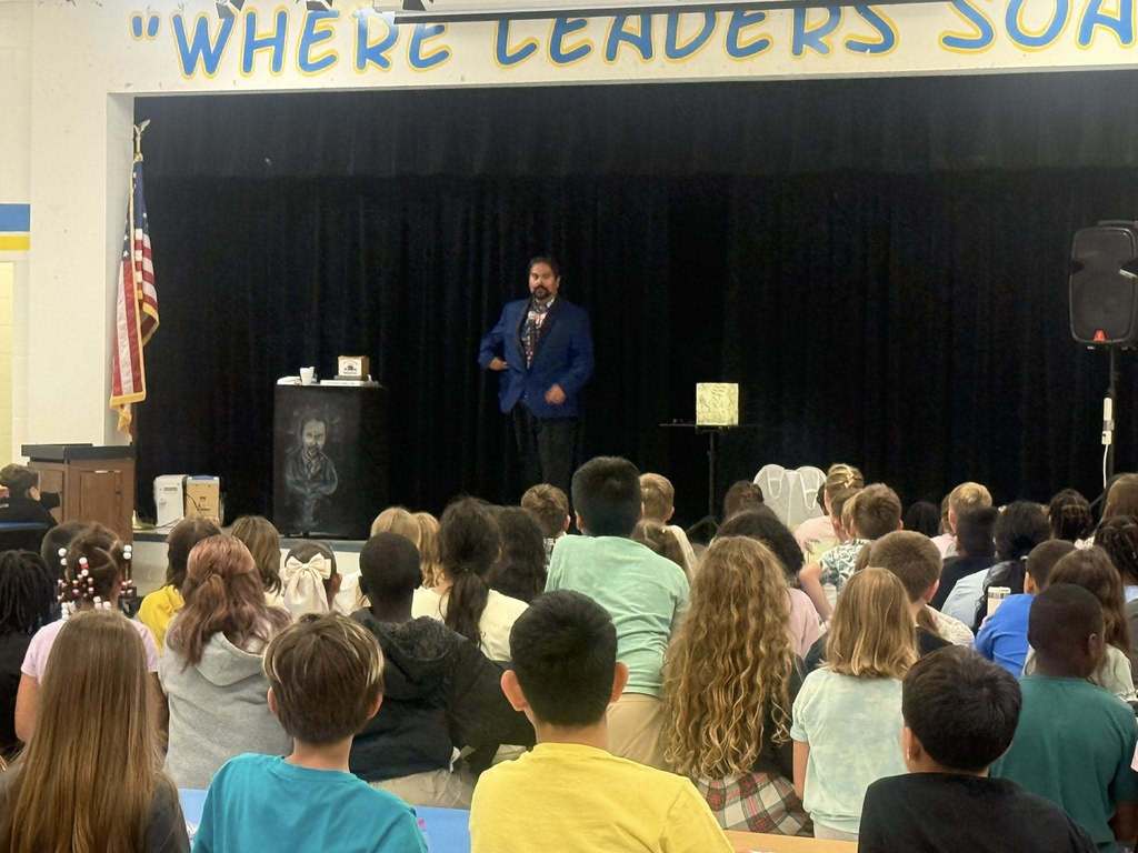 Gemini said A male performer in a blue blazer stands on a stage before a large audience of seated children in a school assembly. Behind him is a black curtain and a sign reading "WHERE LEADERS SOAR." An American flag and a speaker flank the stage. The children's backs face the camera.