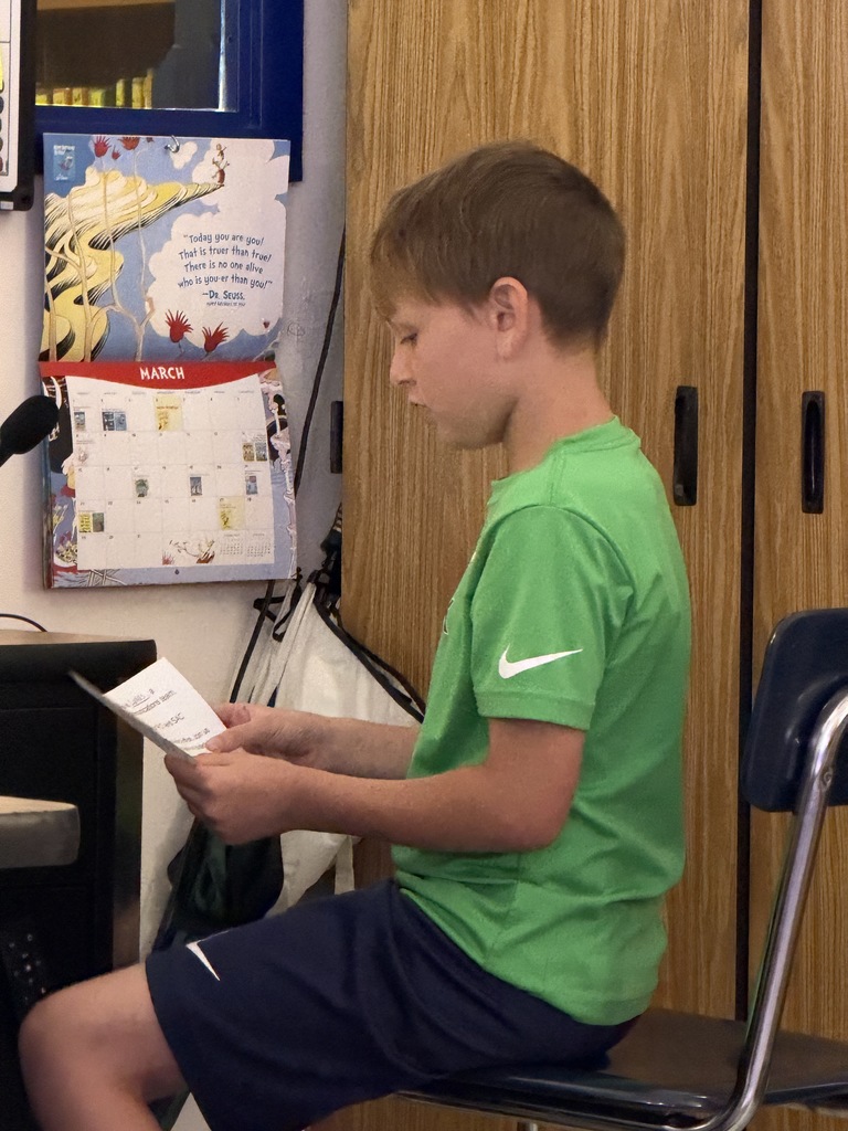 A young boy in a green Nike t-shirt and dark shorts sits on a chair, holding a small white paper with both hands. He is reading the text on the paper intently. In the background, a Dr. Seuss calendar and a microphone are visible.
