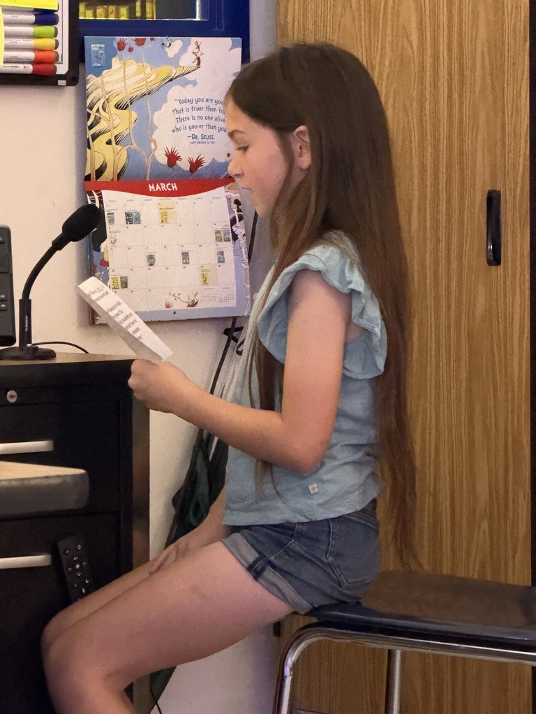 A young girl with long brown hair, wearing a light blue ruffled top and denim shorts, sits on the edge of a chair. She holds a small white paper in her right hand, reading aloud into a desk-mounted microphone positioned in front of her.