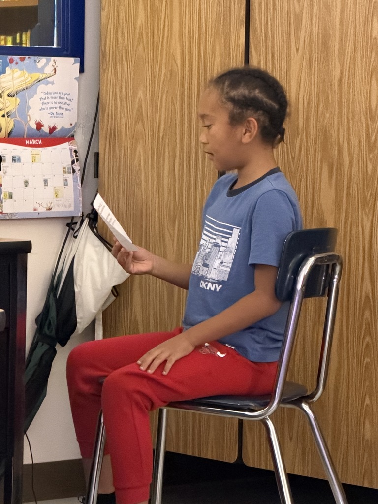 A child with braided hair, wearing a blue DKNY t-shirt and red pants, sits in a metal chair. They are looking down at and reading from a folded white paper held in their left hand against a background of wooden cabinets and a wall calendar.