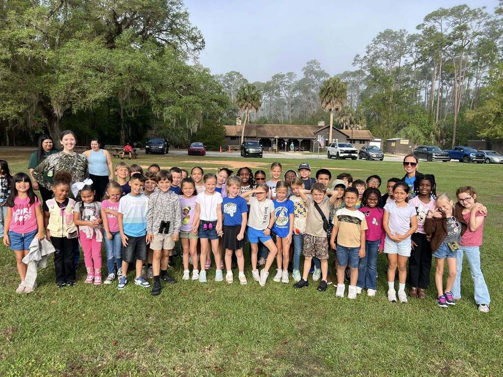 A large group of elementary-aged children and several adults pose for a photo on a grassy field. They are dressed in casual, colorful clothing, some wearing binoculars. In the background, there is a brown wooden building, parked cars, and a dense line of trees under a bright sky.